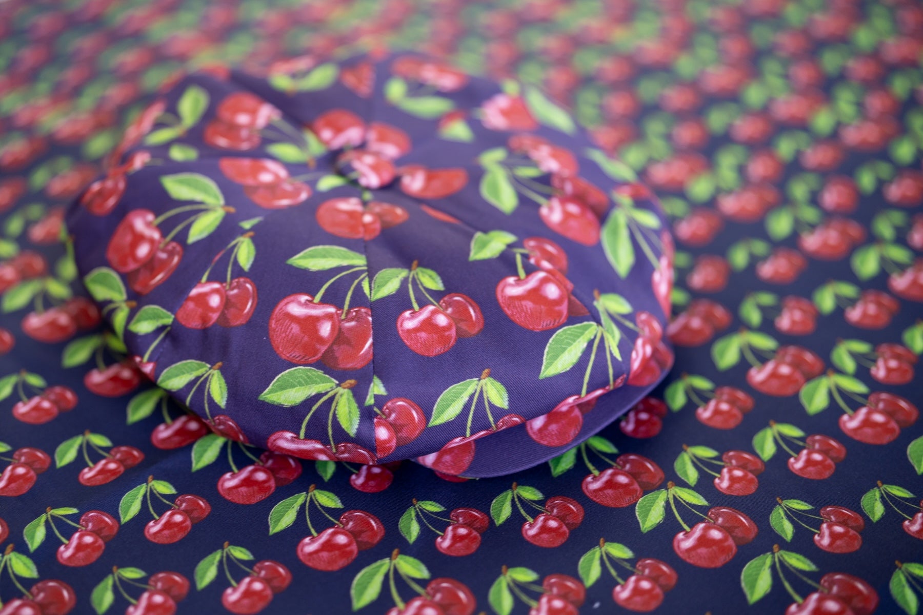 Midnight blue oversized Bakerboy cap with bold exploding cherry print — Cherry Pop hat from the Leigh Francis x Mister Miller collection, featuring curved peak and 70s-style swirl lining. Hat laid on the printed fabric