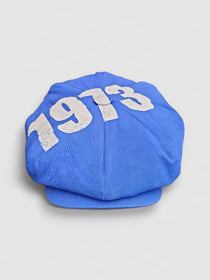 Leigh Francis x Mister Miller 1973 Bakerboy cap — full-crowned cotton twill cap in blue with bold ‘1973’ print featuring a curved snap peak. Front view.
