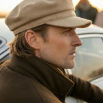 A man in profile wearing a beige newsboy cap and a brown jacket, standing outdoors near a car with a blurred person in the background.