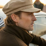 A man in profile wearing a beige newsboy cap and a brown jacket, standing outdoors near a car with a blurred person in the background.