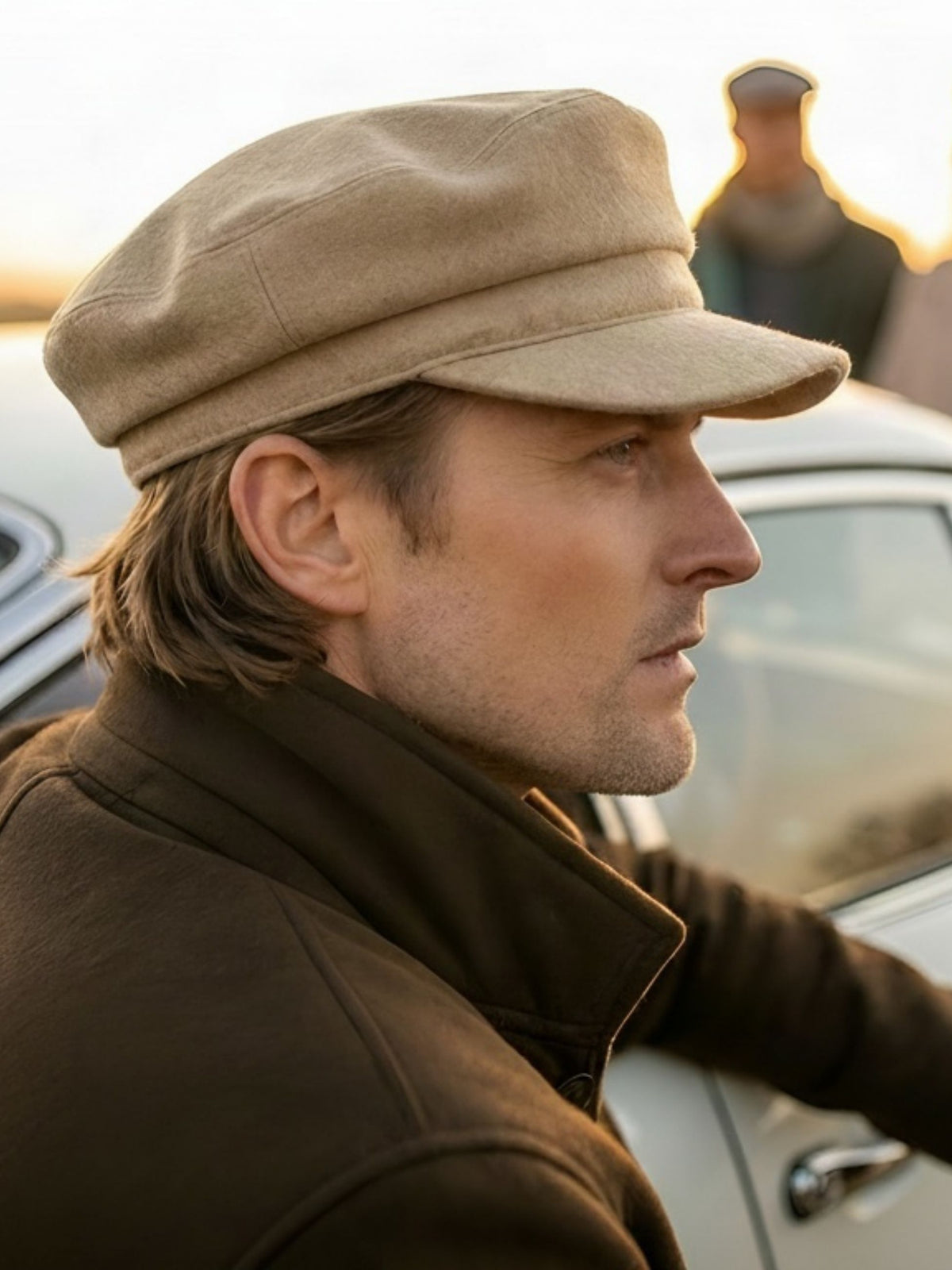 A man in profile wearing a beige newsboy cap and a brown jacket, standing outdoors near a car with a blurred person in the background.