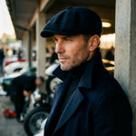 A man wearing a dark navy blue flat cap and matching dark coat, standing in a covered outdoor area with cars and concrete pillars in the background.