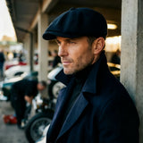 A man wearing a dark navy blue flat cap and matching dark coat, standing in a covered outdoor area with cars and concrete pillars in the background.