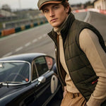 A man wearing a green flat cap, dark quilted vest, beige turtleneck, and brown trousers stands beside a vintage black car on a racetrack.