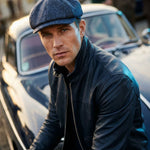 A man wearing a dark blue textured flat cap and a black leather jacket, posing in front of a vintage car.