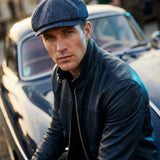 A man wearing a dark blue textured flat cap and a black leather jacket, posing in front of a vintage car.