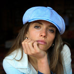 A woman wearing a Bakerboy cap in light blue Irish linen herringbone — Riley Skies by Mister Miller, a breathable summer hat handmade in Britain. 