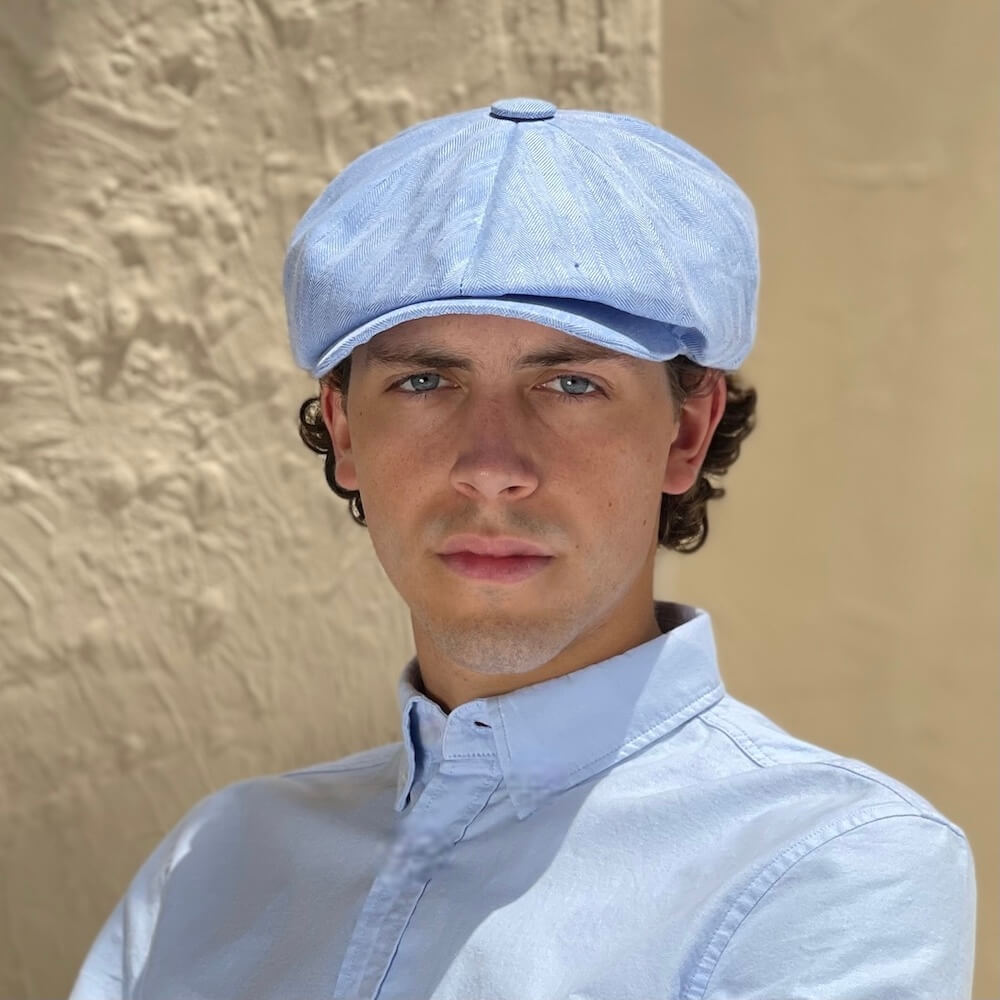 A man wearing a Bakerboy cap in light blue Irish linen herringbone — Riley Skies by Mister Miller, a breathable summer hat handmade in Britain. 