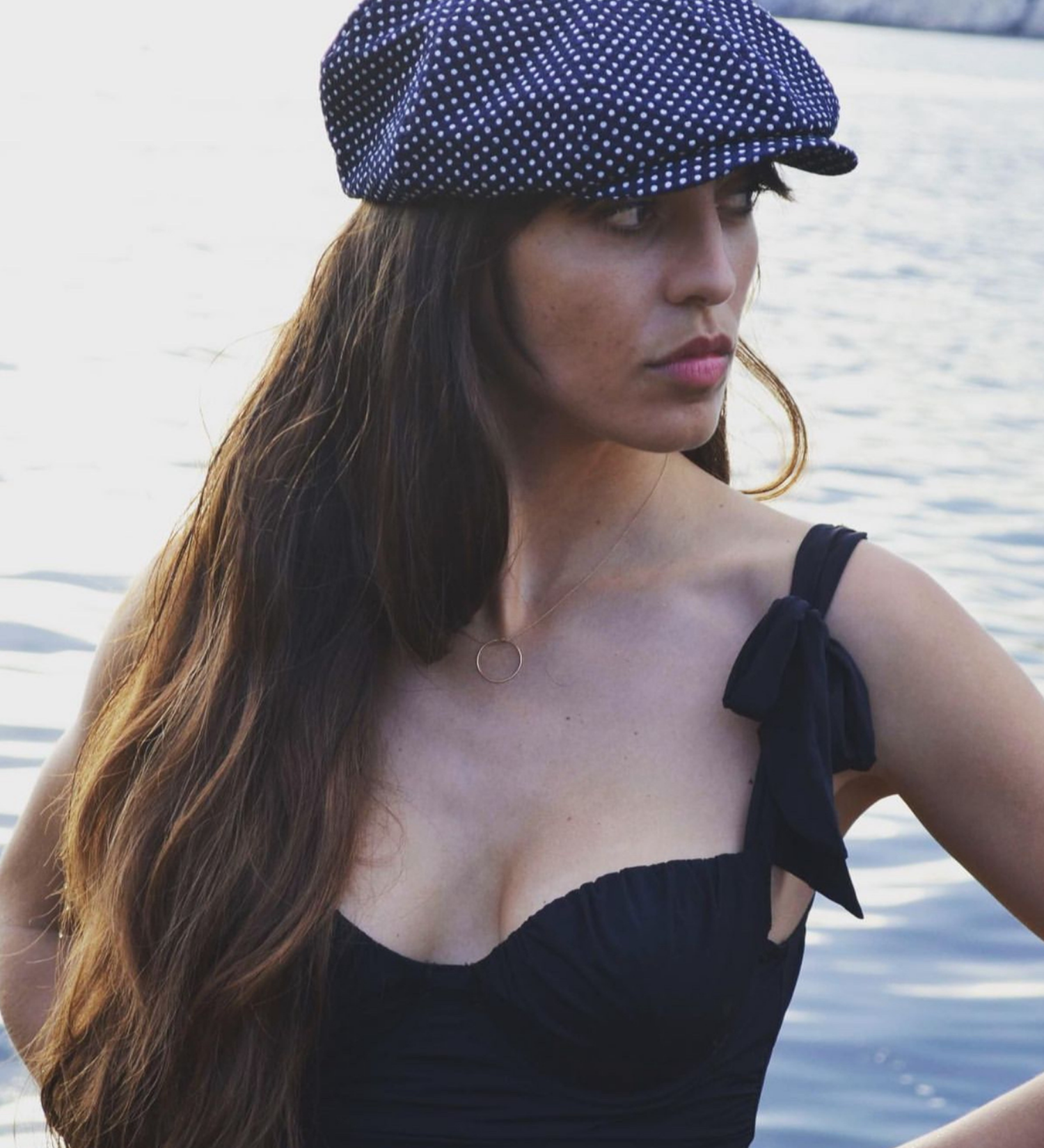 Woman wearing a navy polka dot cap and black dress by a body of water.