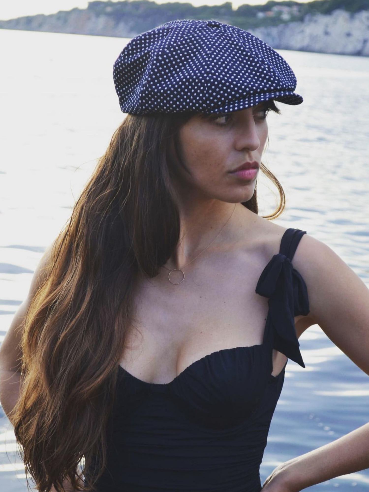 Woman wearing a navy polka dot cap and black dress by a body of water.