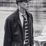 Man wearing a jacket, tie, and cap standing by a water