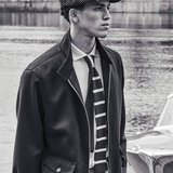Man wearing a jacket, tie, and cap standing by a water