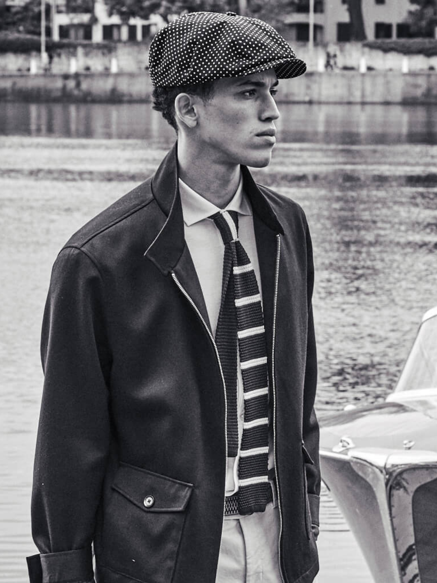Man wearing a jacket, tie, and cap standing by a water