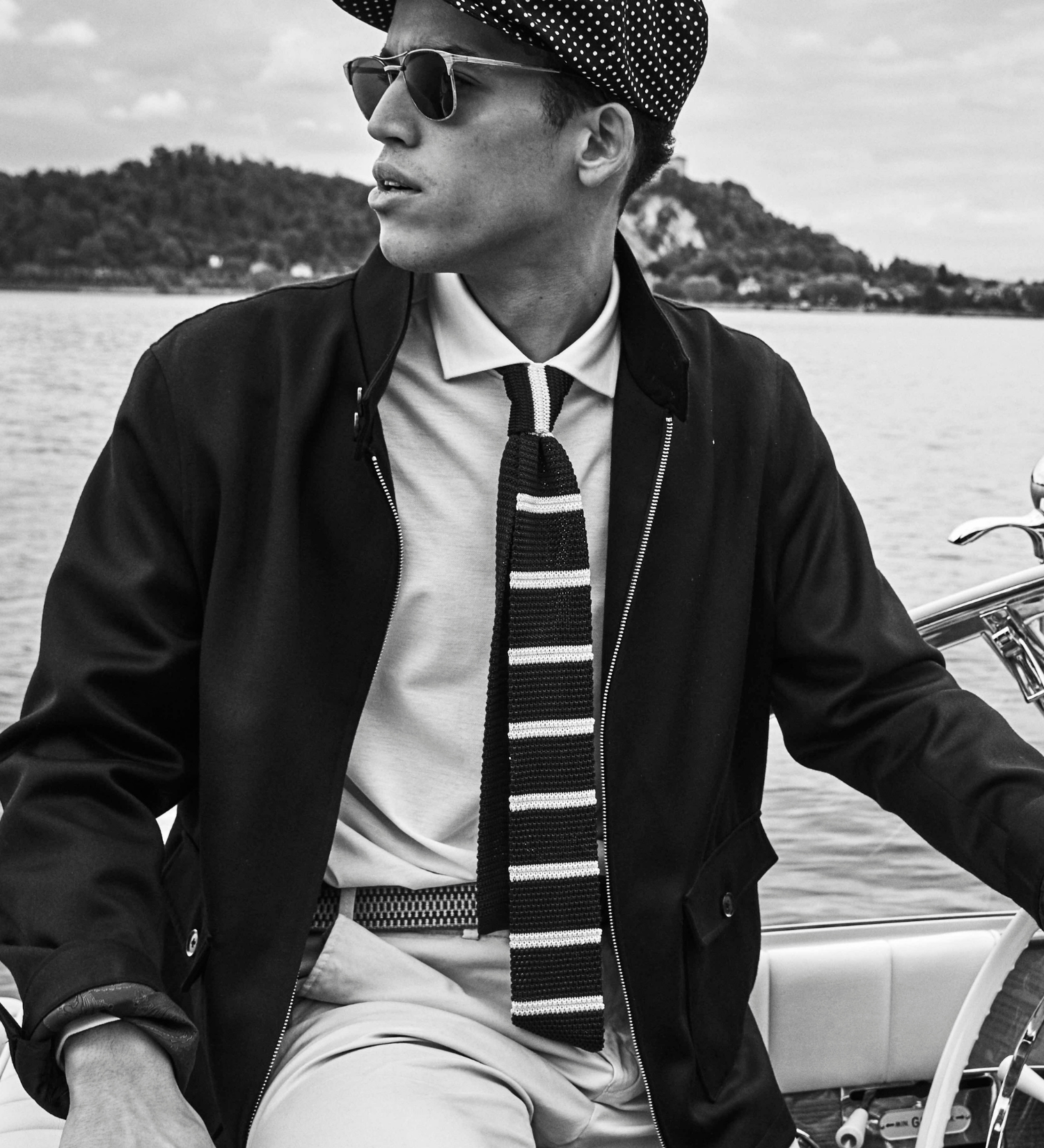 Man wearing a cap, sunglasses, and a striped tie on a boat with a scenic background
