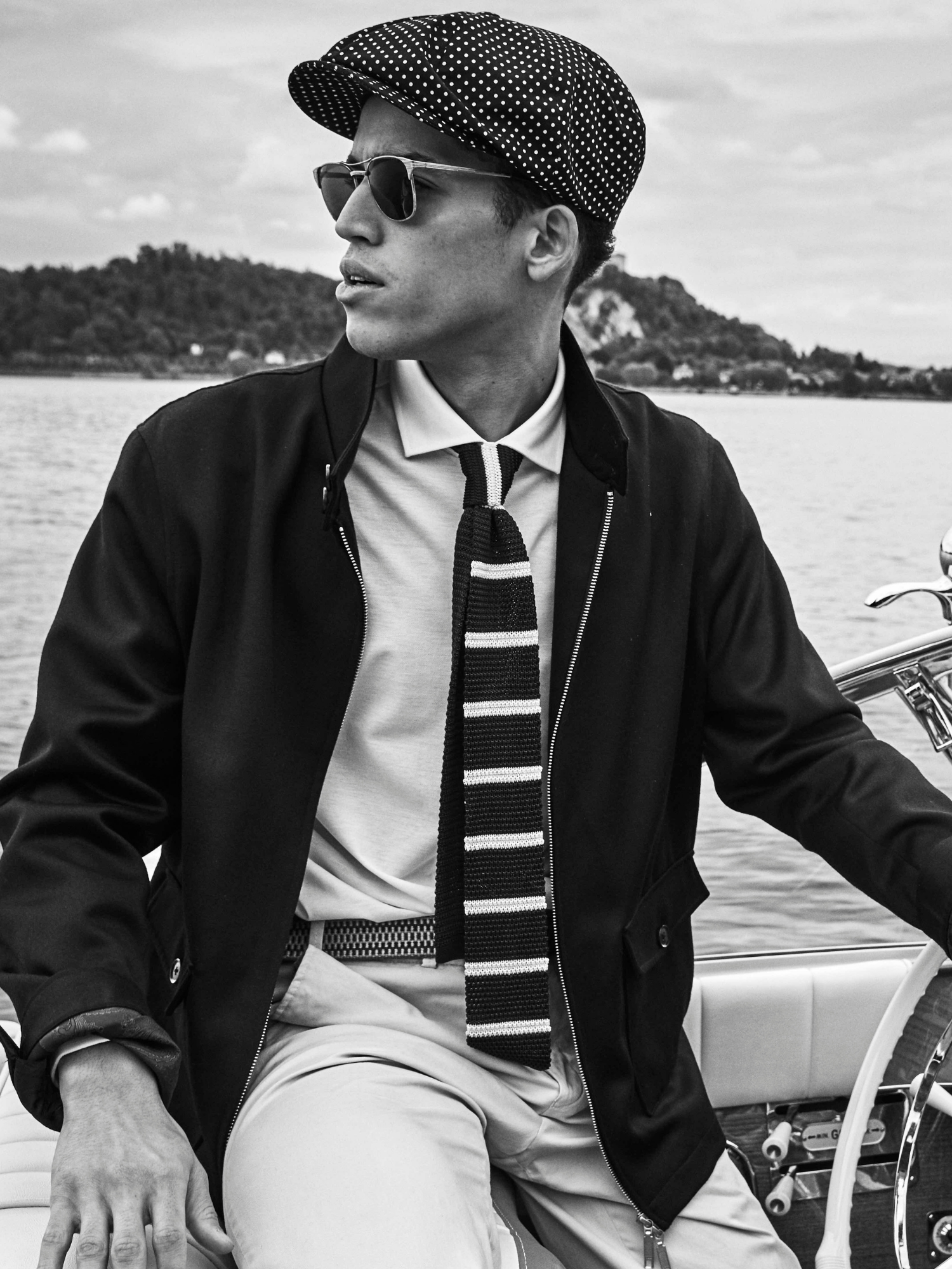 Man wearing a cap, sunglasses, and a striped tie on a boat with a scenic background