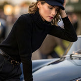 A woman wearing a black bakerboy cap with dark turtleneck sweater, matching dark trousers, leans against the hood of a classic porsche in an outdoor setting.