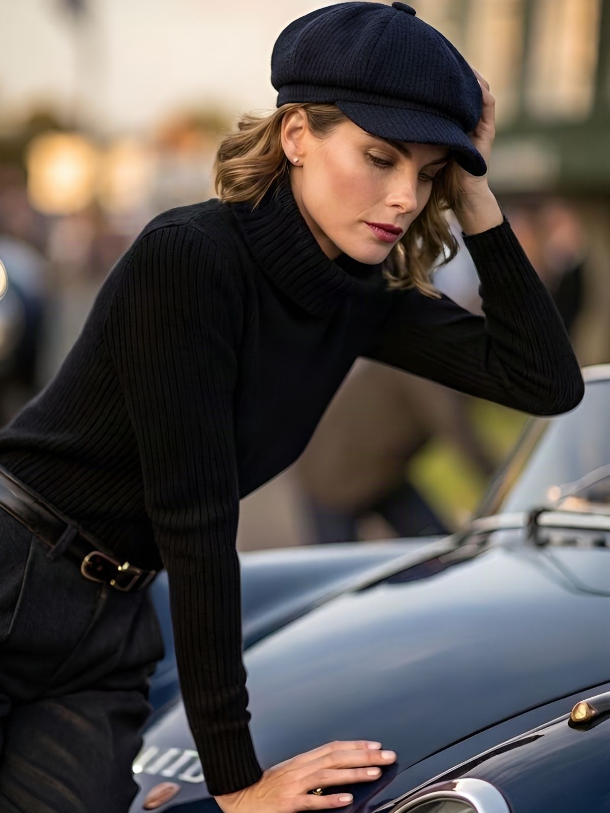 A woman wearing a black bakerboy cap with dark turtleneck sweater, matching dark trousers, leans against the hood of a classic porsche in an outdoor setting.